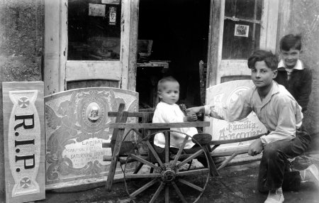 Retrat de tres nens davant el taller del carrer de Santa Magdalena, 10, a Olot; en primer terme Sadurní Brunet Forasté i damunt del carretó el seu germà Jordi, 1928 (ACGAX. Fons Sadurní Brunet Pi. Autor: Sadurní Brunet)