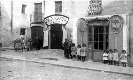 Vista frontal de la farmàcia Pujol i la xocolateria Pi, a Tortellà, 1917, (ACGAX. Fons Sadurní Brunet Pi. Autor: Sadurní Brunet)
